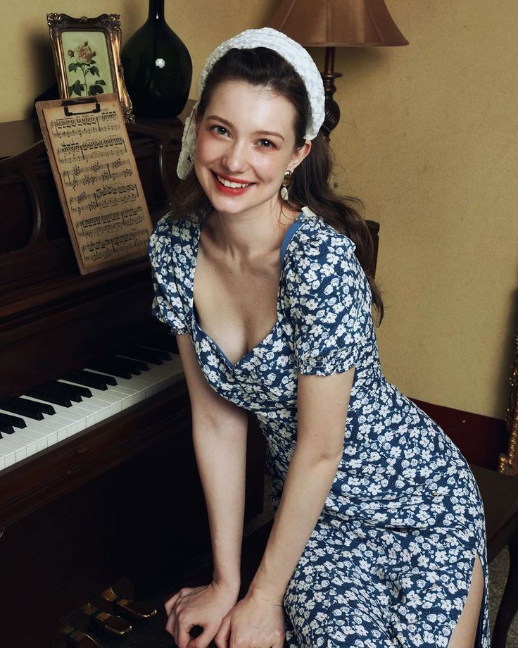 Woman in a blue floral dress and white headband posing by a piano