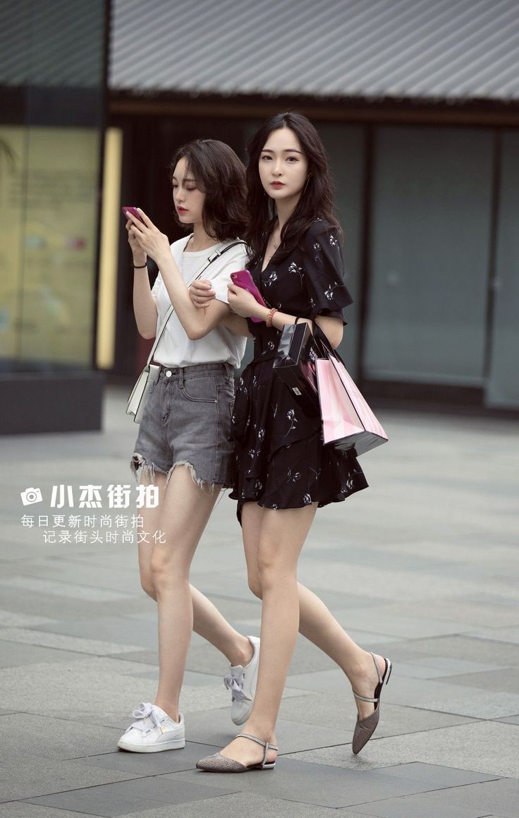 Two women walking on a street, one in a black dress and the other in denim shorts