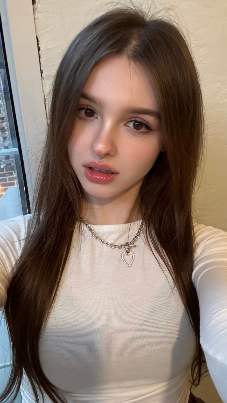 Woman with long brown hair wearing a white top and heart necklace indoors