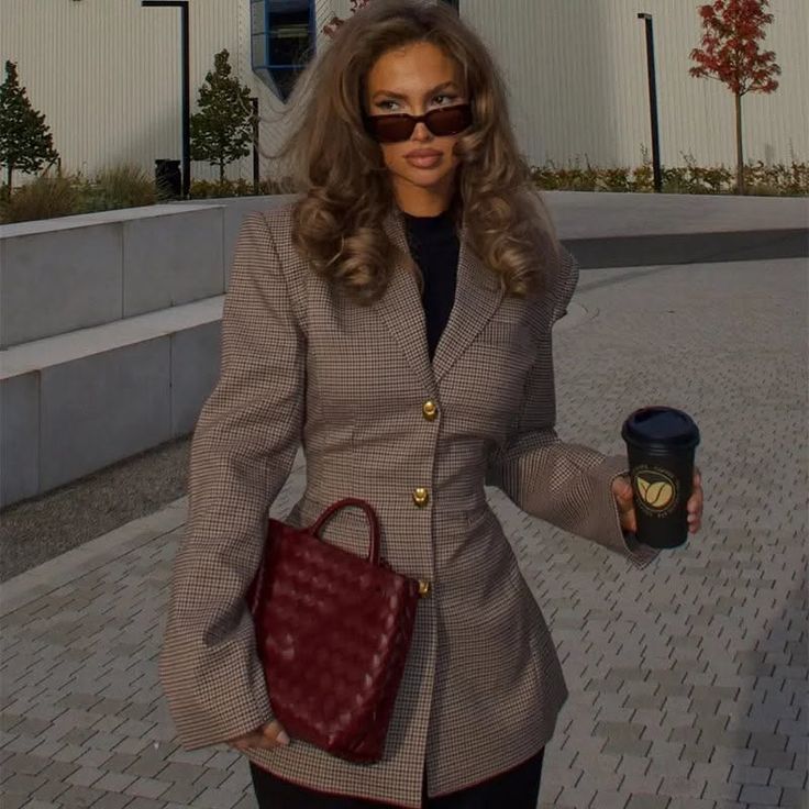 Woman with long hair wearing a blazer and sunglasses, holding a red bag and coffee