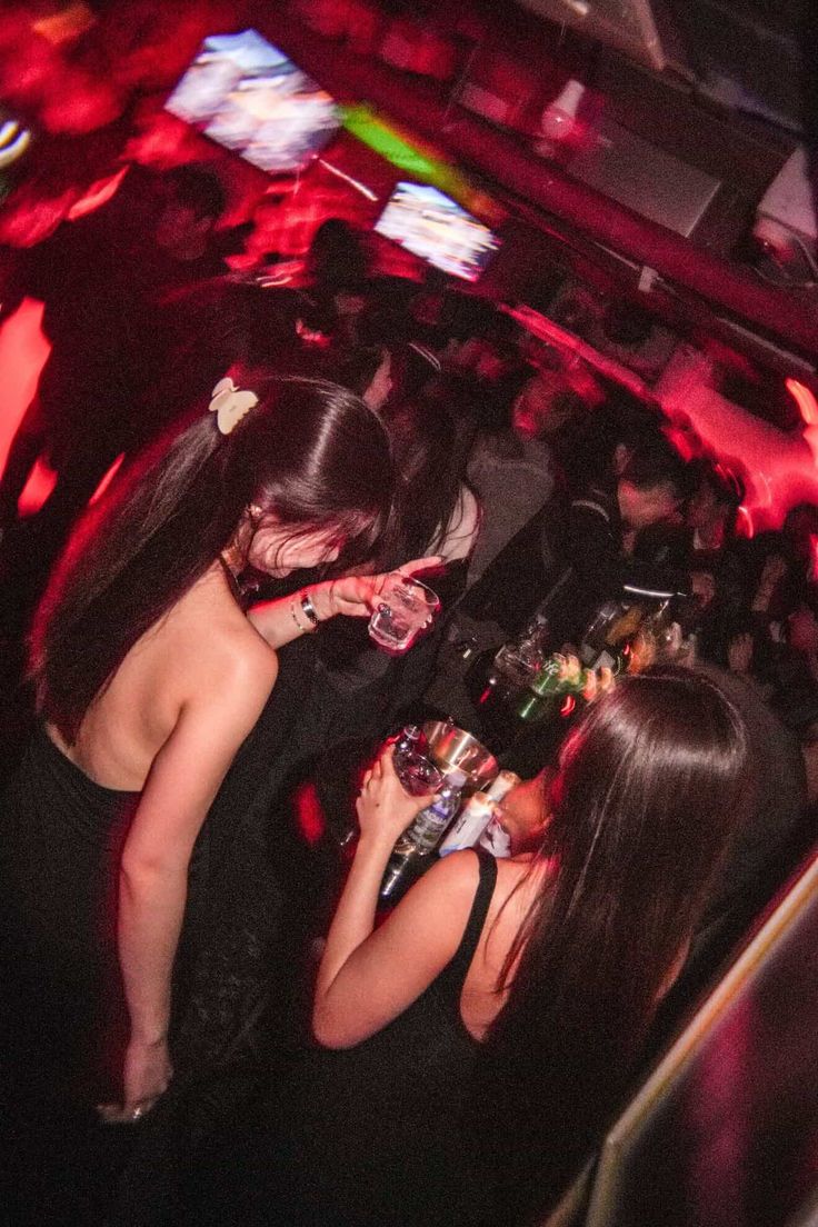 Two women in black dresses drinking in a dimly lit nightclub with red lighting