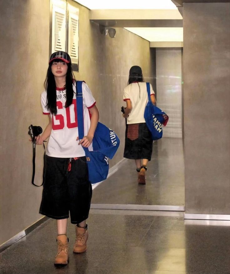 Person in a t-shirt, baggy shorts, and boots holding a camera in a hallway