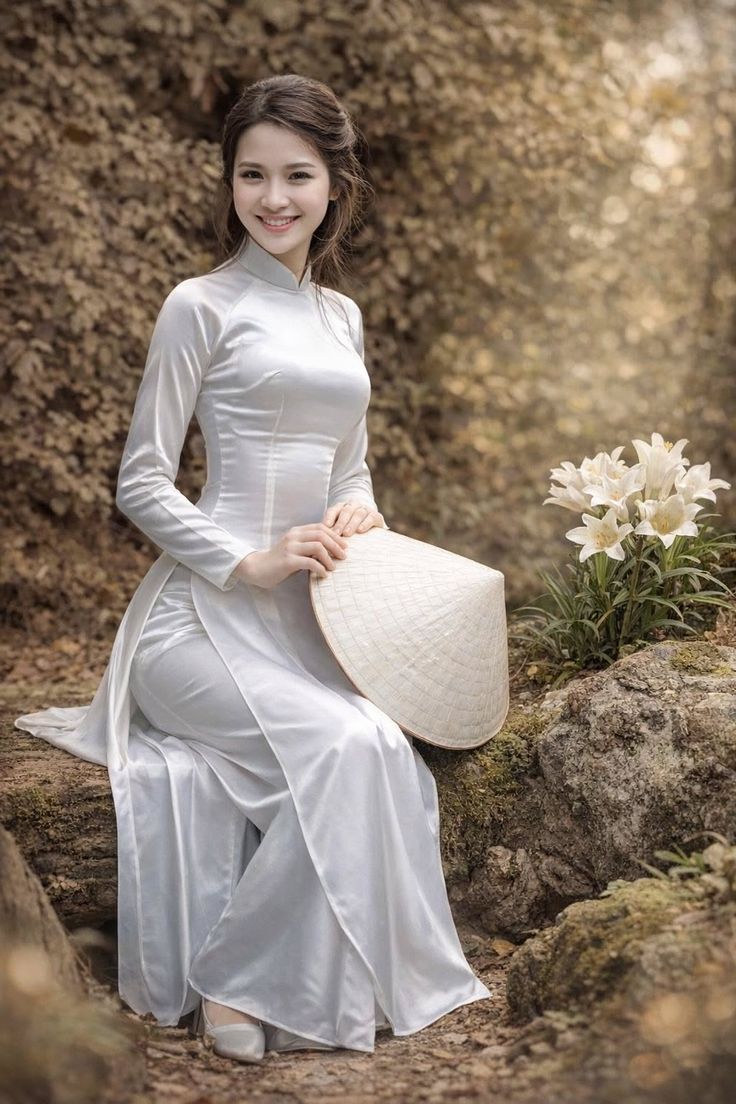 Woman in a white ao dai and conical hat sitting outdoors with white flowers