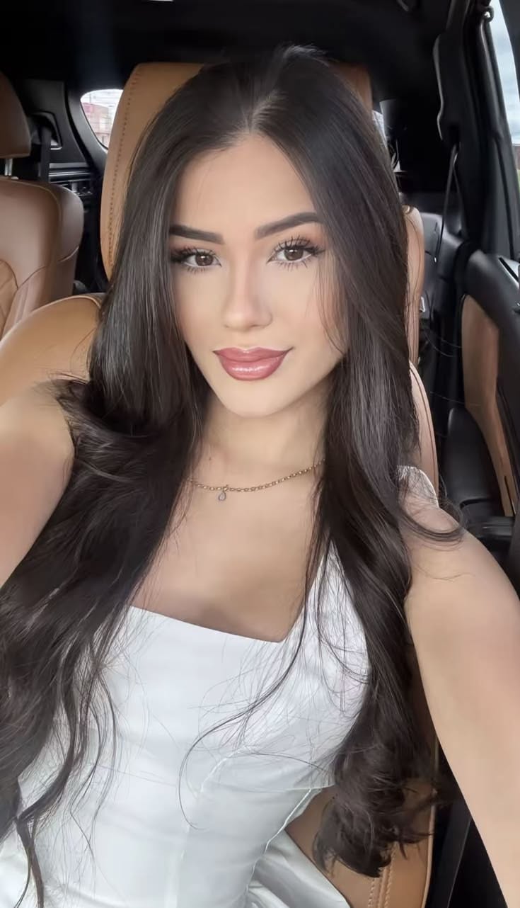 Woman with long dark hair wearing a white top and necklace in a car