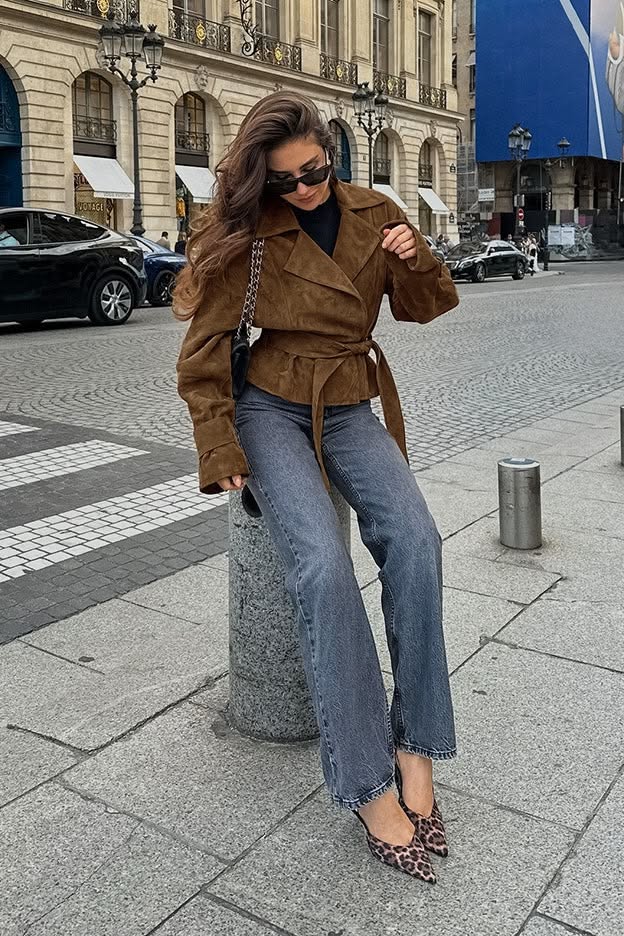 Woman in a brown suede jacket and jeans sitting on a stone pillar in a city