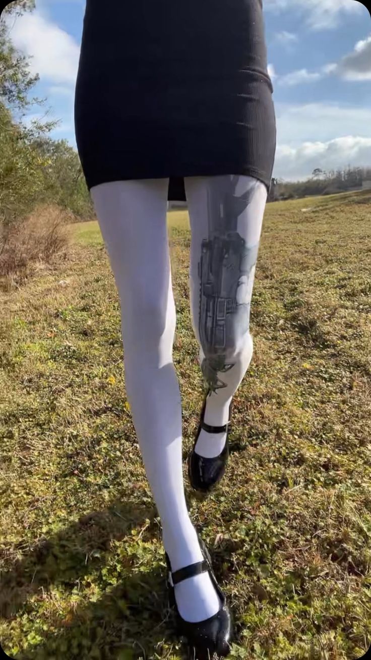 Person in a black dress and white tights with a leg tattoo walking on grass