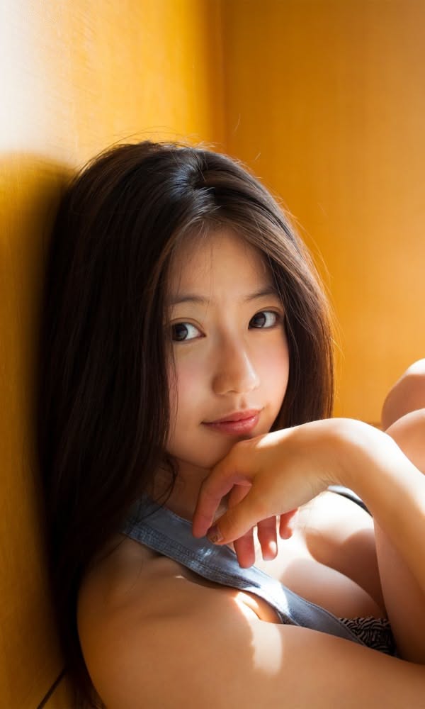 Woman with long black hair wearing a denim top, leaning against a warm-toned wall