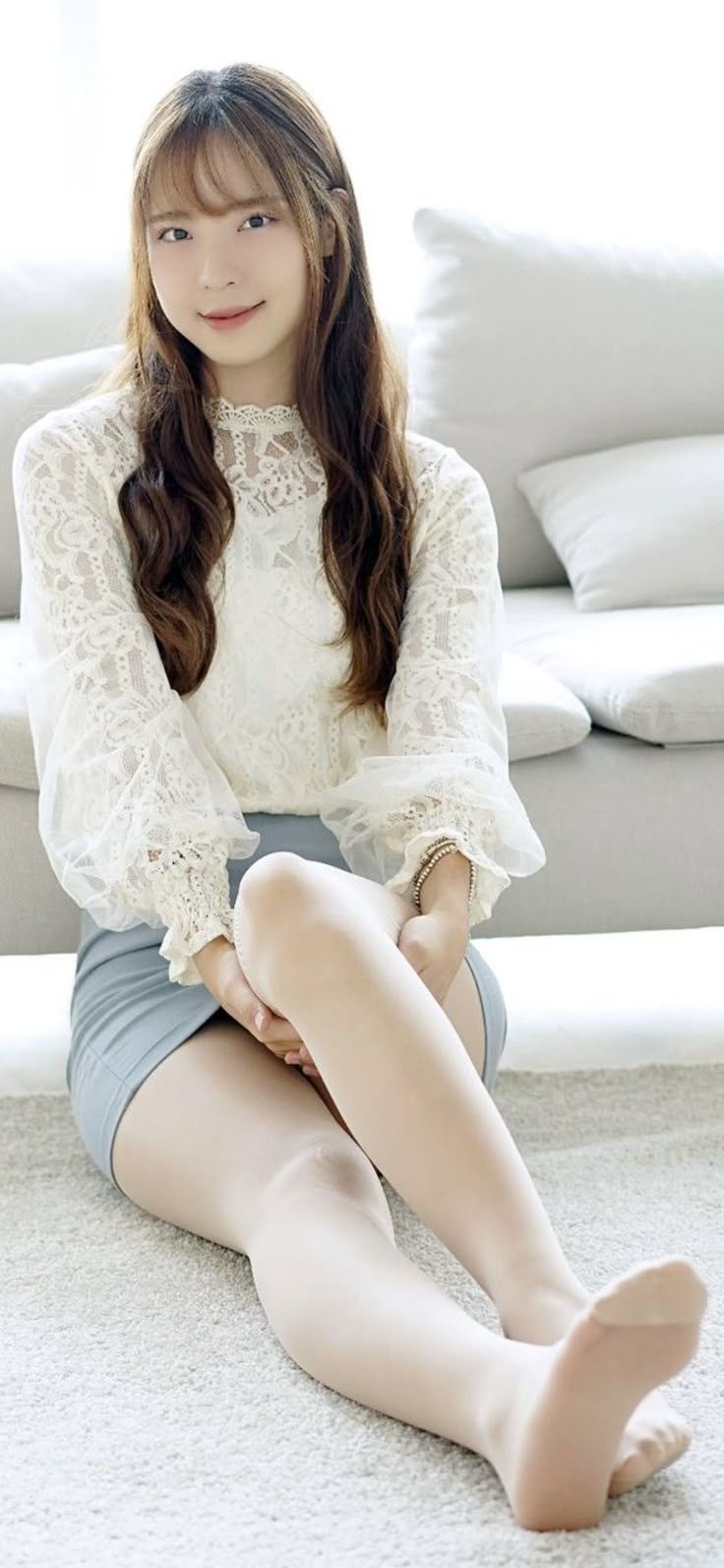 Woman with long hair wearing a white lace blouse and mini skirt, sitting indoors