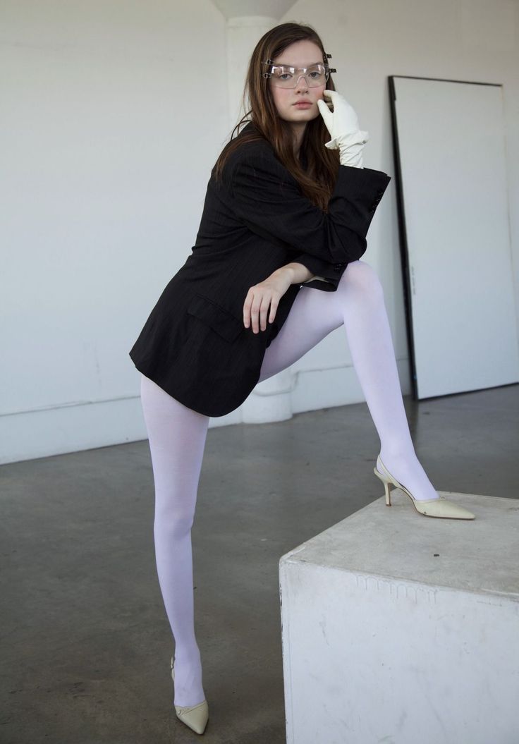 Woman in a black blazer, white tights, and clear glasses posing indoors