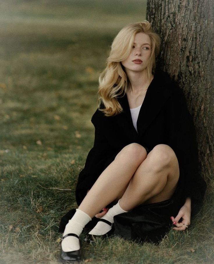 Blonde woman in a black coat and white socks sitting on grass next to a tree