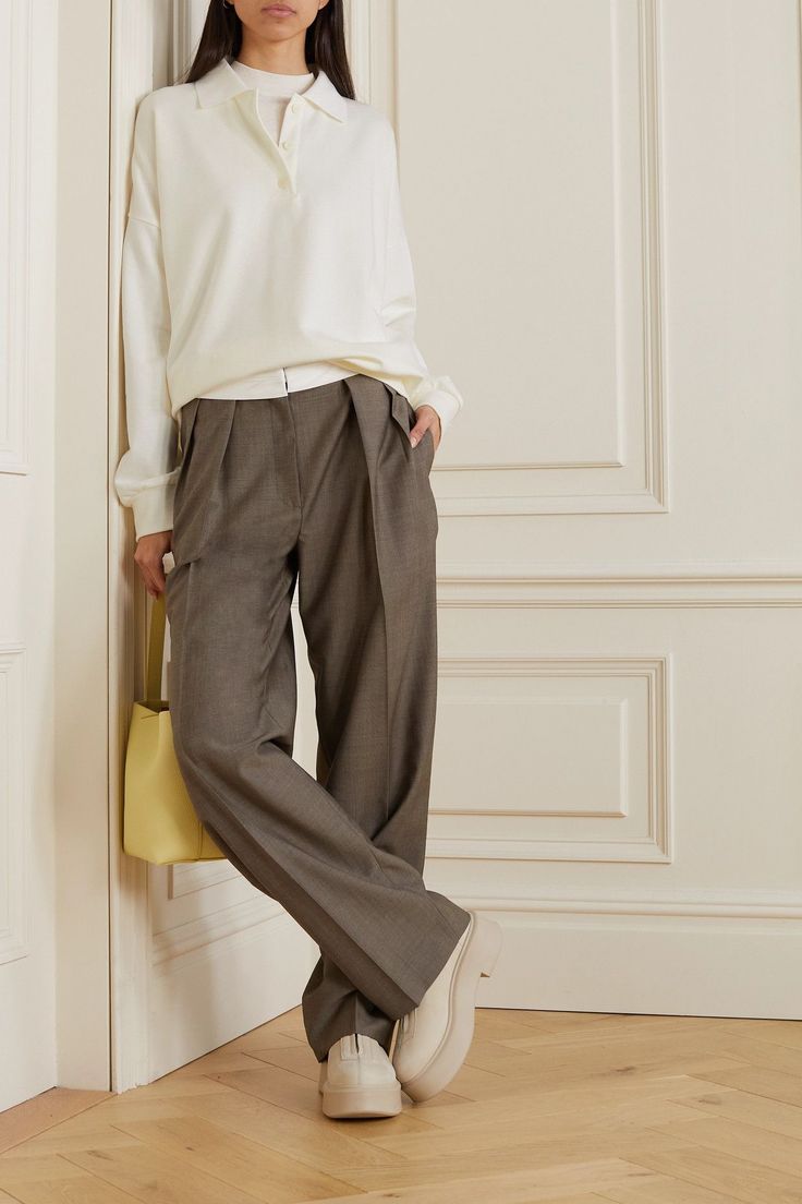 Woman in a white long sleeve polo and wide leg pants leaning against a wall