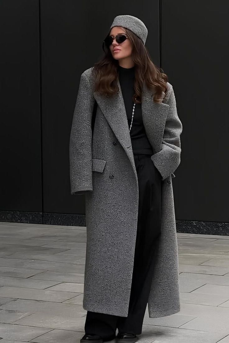 Woman in a long gray coat, black pants, and a hat standing against a dark background