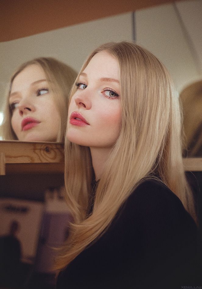 Blonde woman with long hair looking at the camera, with her reflection in a mirror