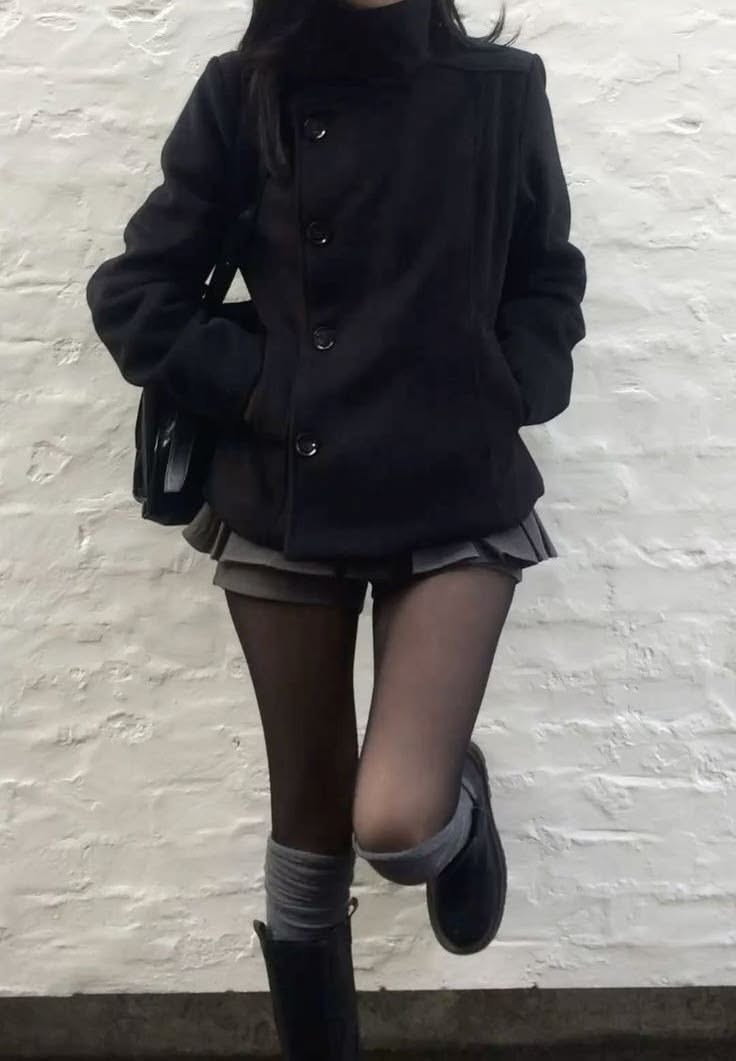 Person in a black coat, pleated skirt, and tights leaning against a white brick wall