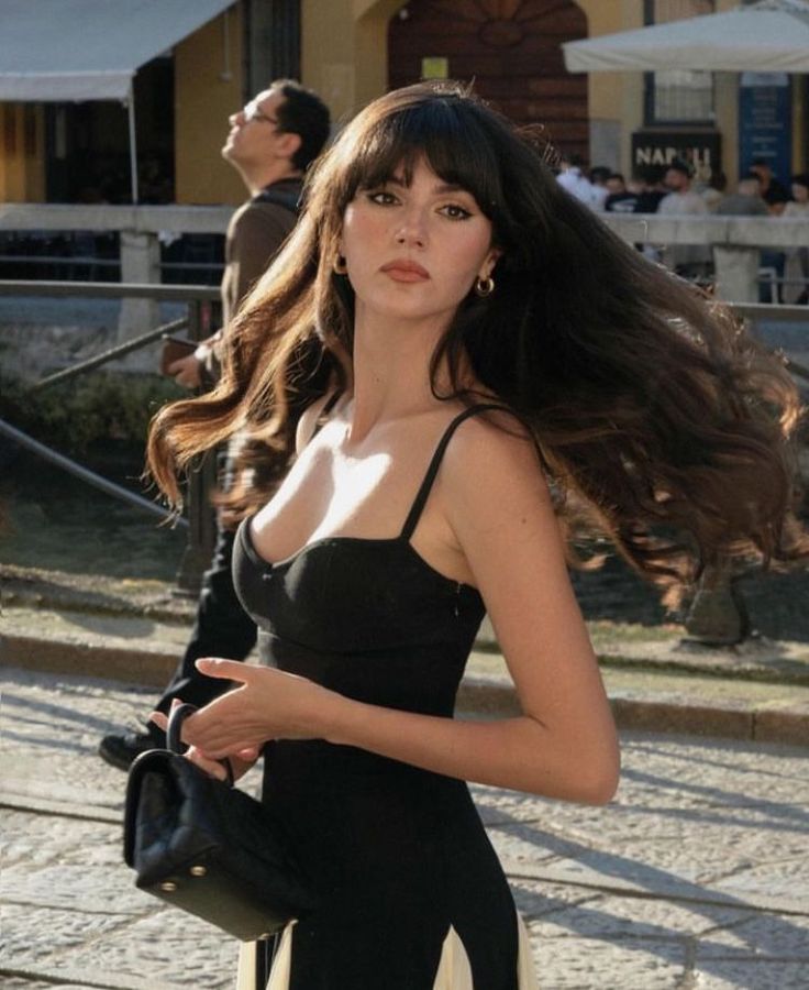 Woman in a black dress with long hair and bangs holding a handbag outdoors