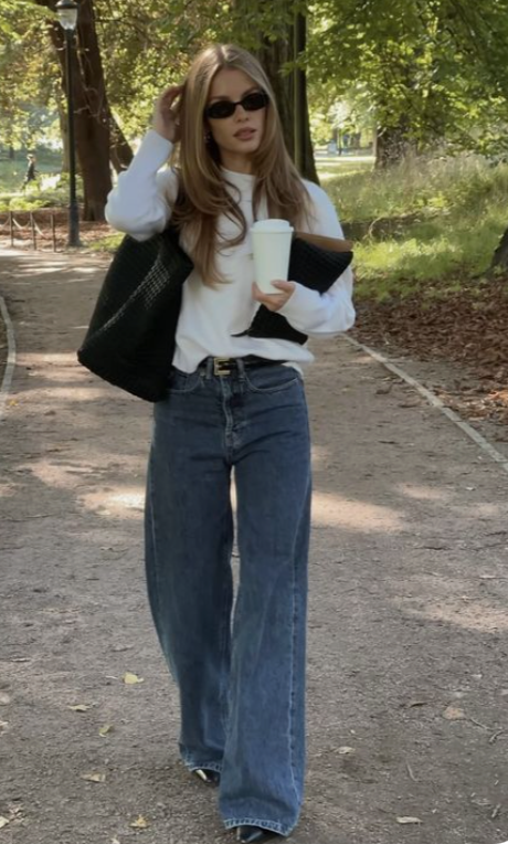 Woman in sunglasses and wide leg jeans walking in a park with a coffee cup