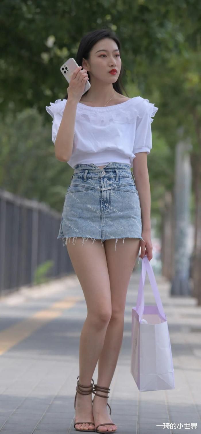 Woman in a white top and denim skirt holding a phone and a bag outdoors