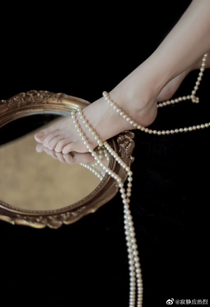 Bare feet adorned with pearls resting on an ornate mirror against a dark background