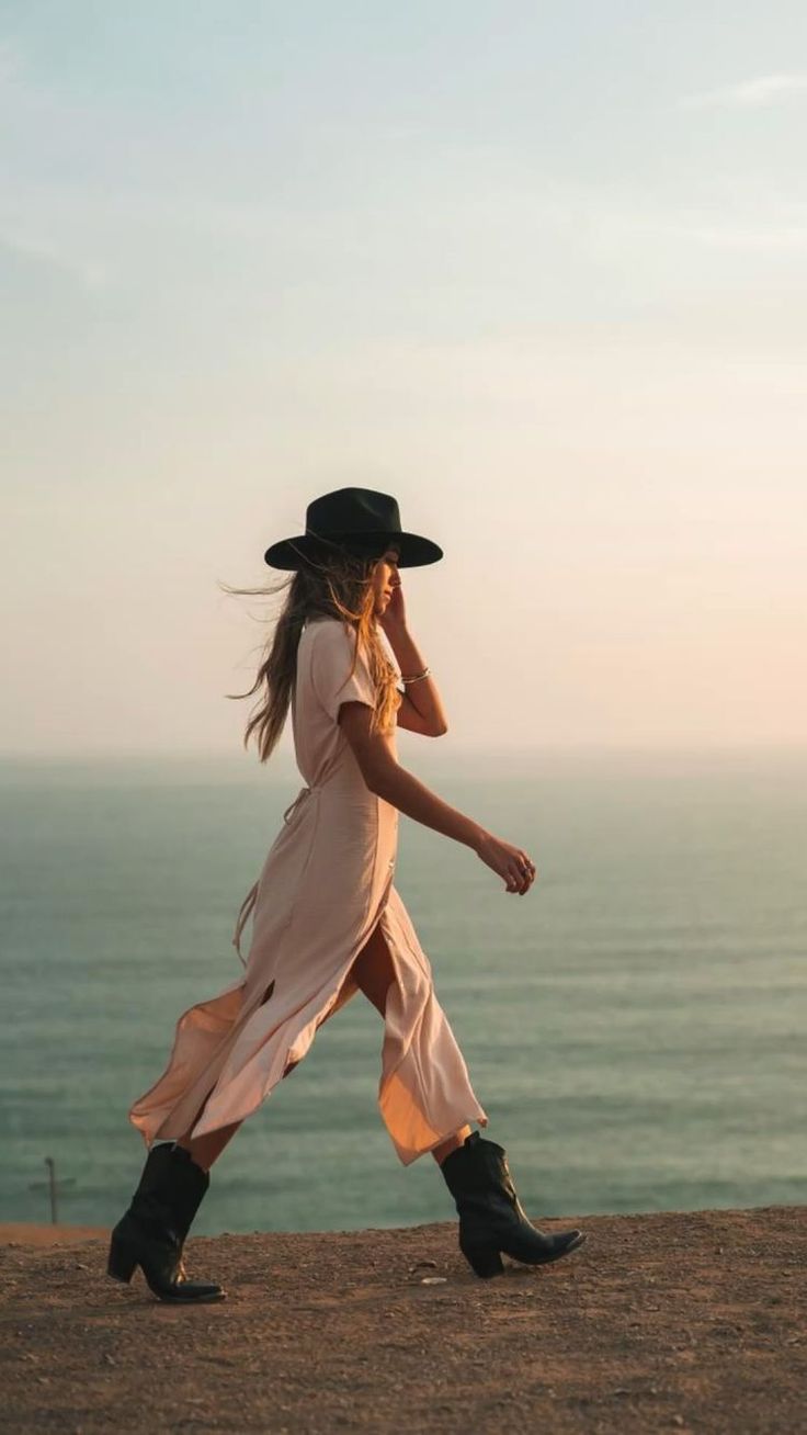 Woman in a long dress and hat walking along a cliff overlooking the ocean at golden hour
