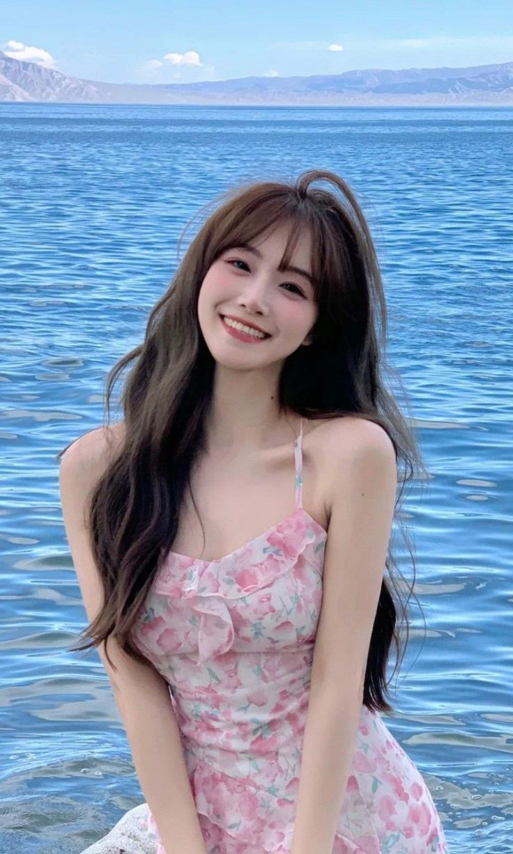 Woman in a pink floral dress smiling by blue water with mountains in the background