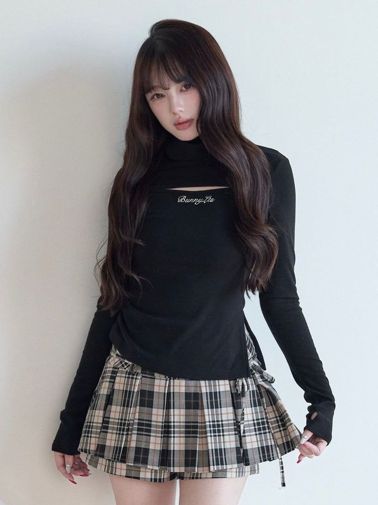 Woman with long dark hair wearing a black top and plaid skirt indoors