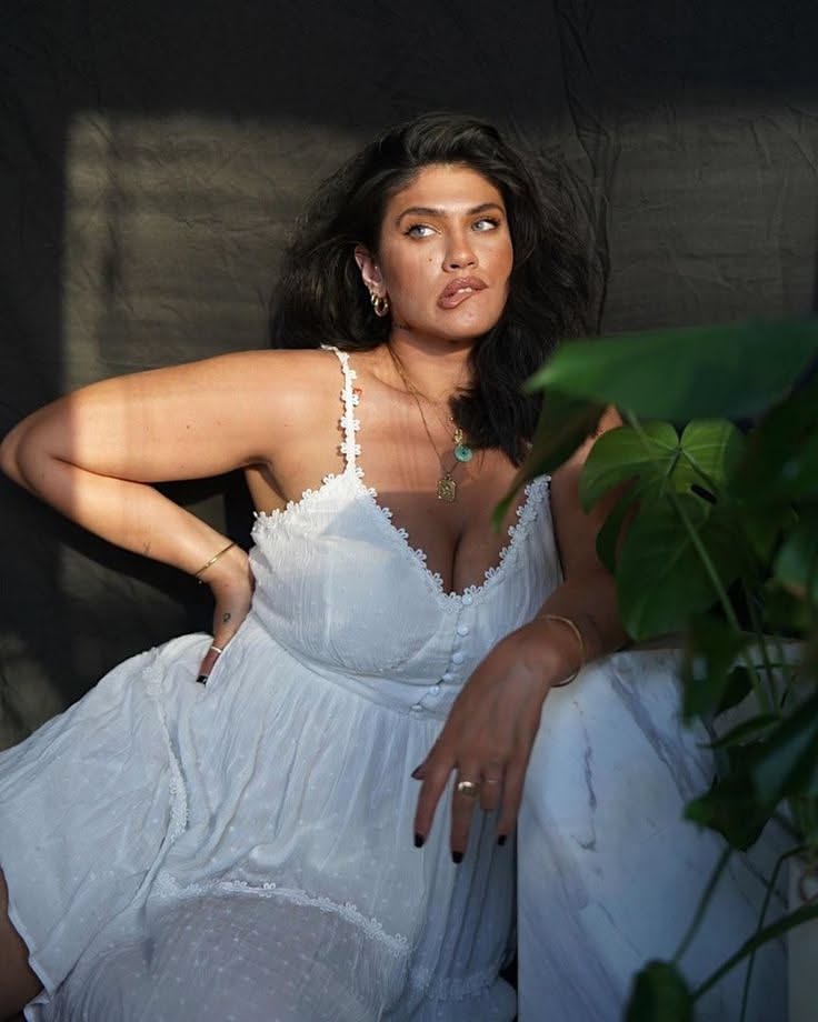 Woman in a white dress with cleavage and jewelry, posing indoors with dramatic lighting
