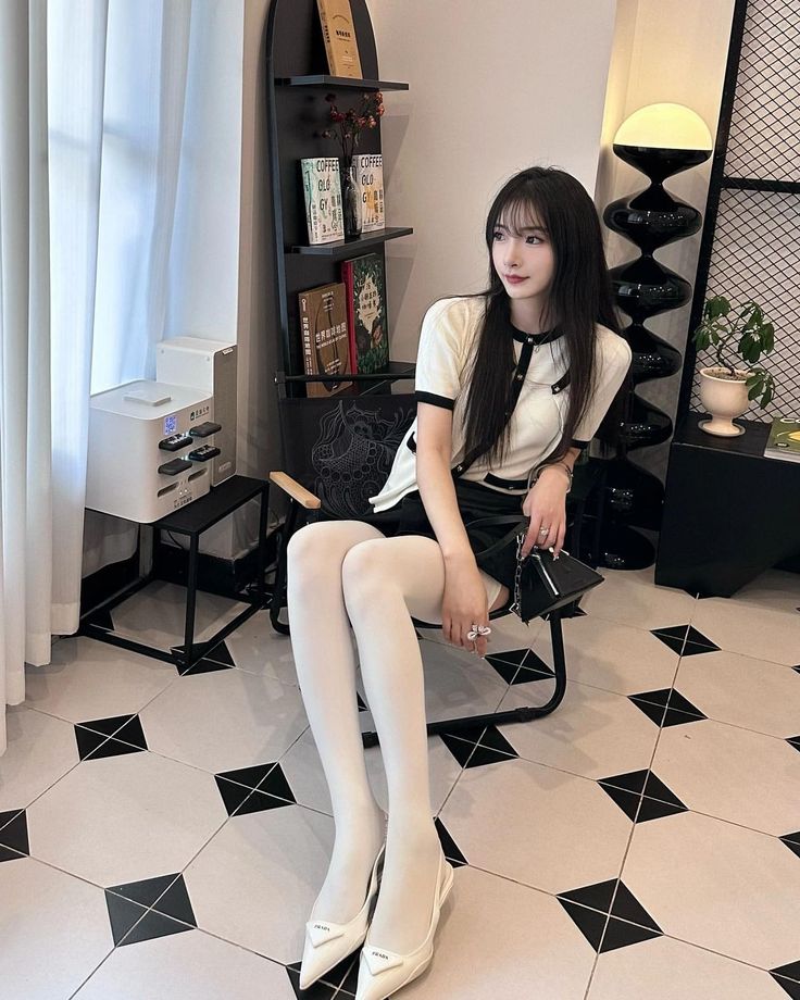 Woman with long black hair wearing a white top and black skirt with white tights, sitting indoors