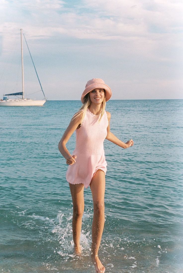 Woman in a pink romper and bucket hat running in the ocean with a sailboat in the background