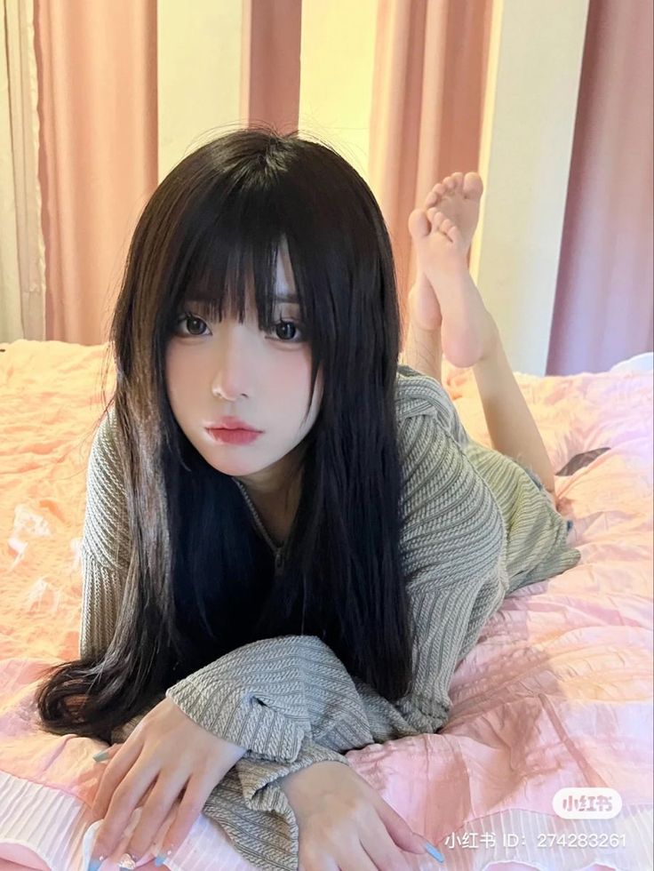 Woman with long black hair and bangs lying on a pink bed in a knitted top