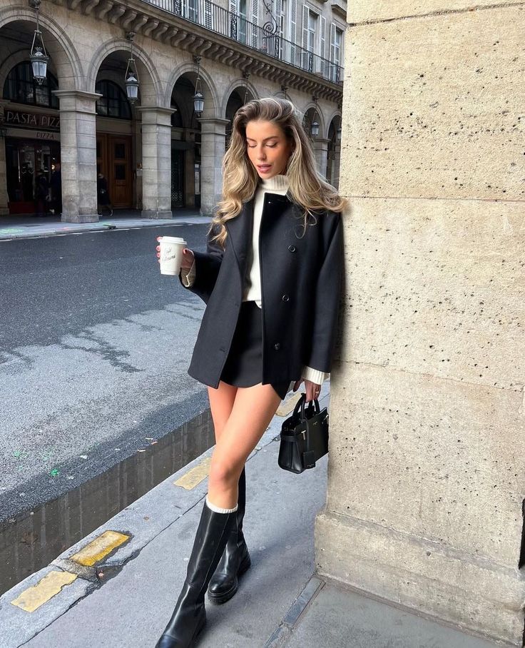 Woman in a black coat and mini skirt holding a coffee cup on a city street