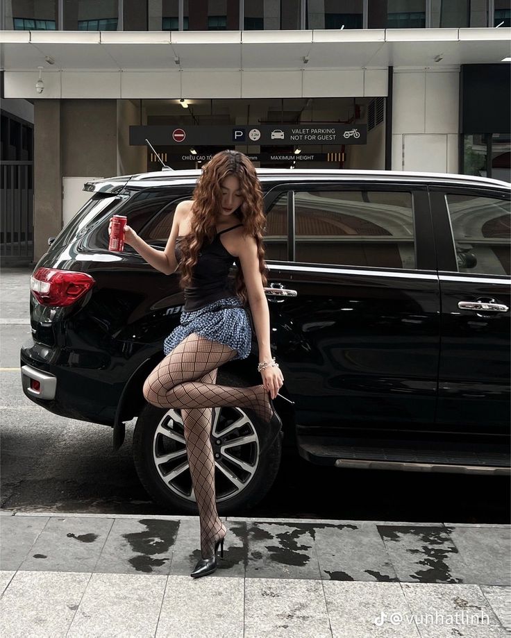 Woman in fishnet stockings and a mini skirt posing next to a black car