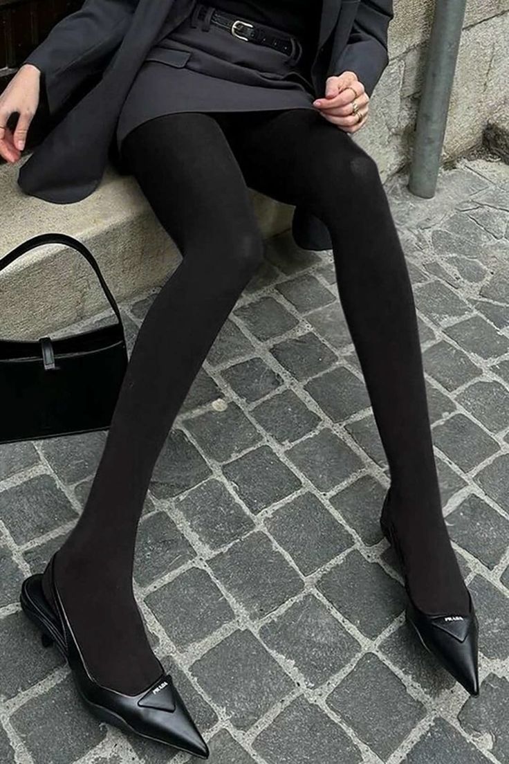 Person in black tights, blazer, and mini skirt sitting on a stone ledge