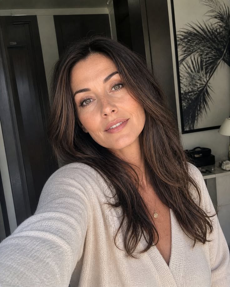 Woman with long brown hair wearing a cream sweater and gold necklace indoors