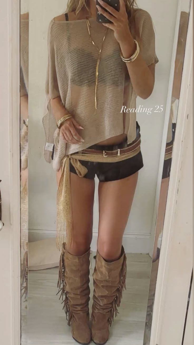 Woman in a sheer top, black shorts, and fringe boots taking a mirror selfie