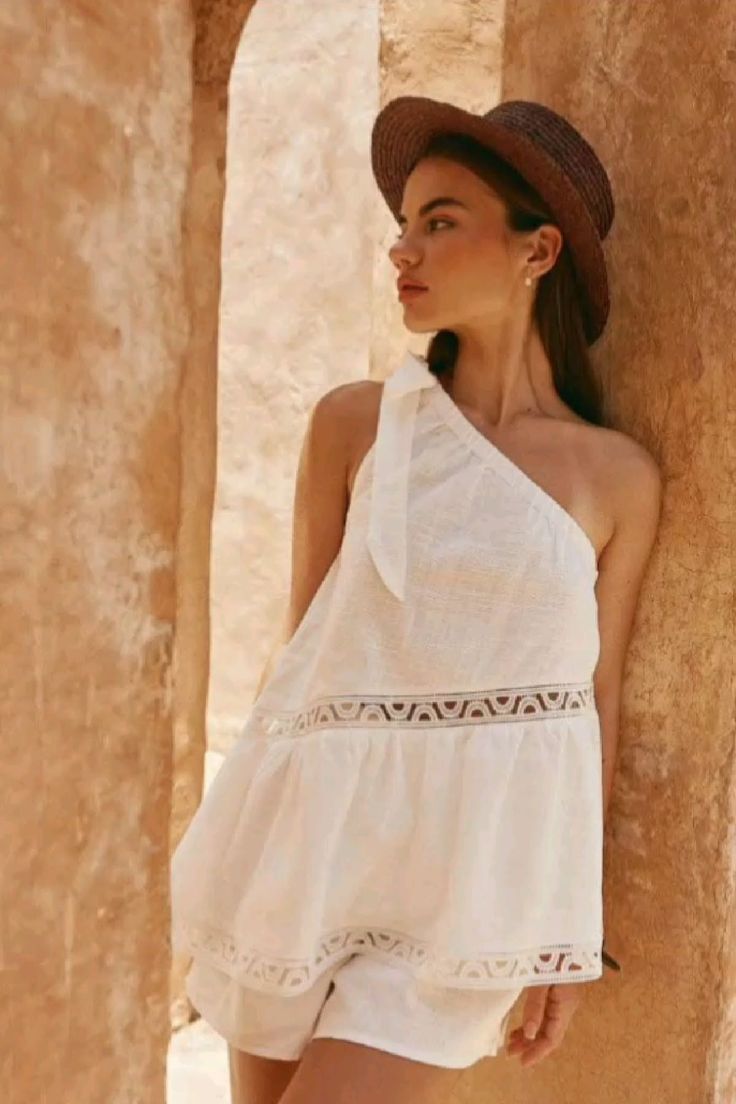 Woman in a white one-shoulder top and straw hat leaning against a wall