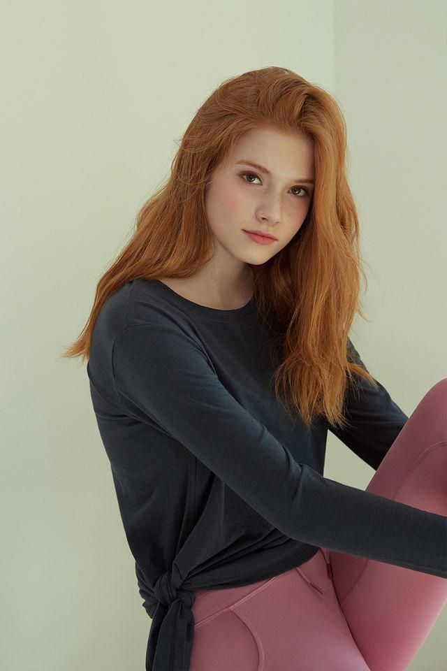 Woman with long red hair and freckles wearing a dark long sleeve shirt and pink leggings
