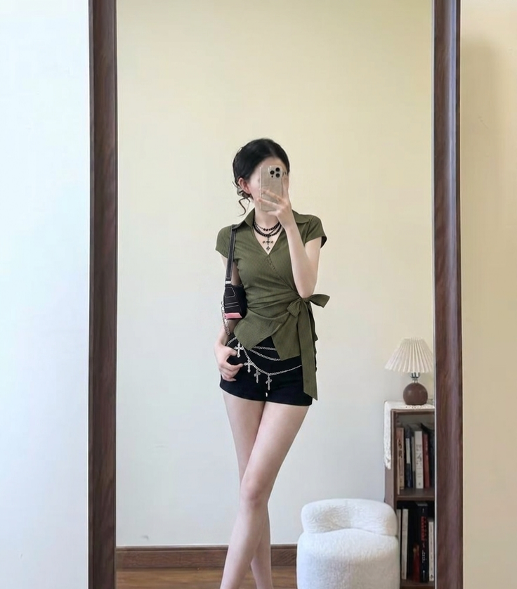 Woman in a green wrap top and black shorts taking a mirror selfie