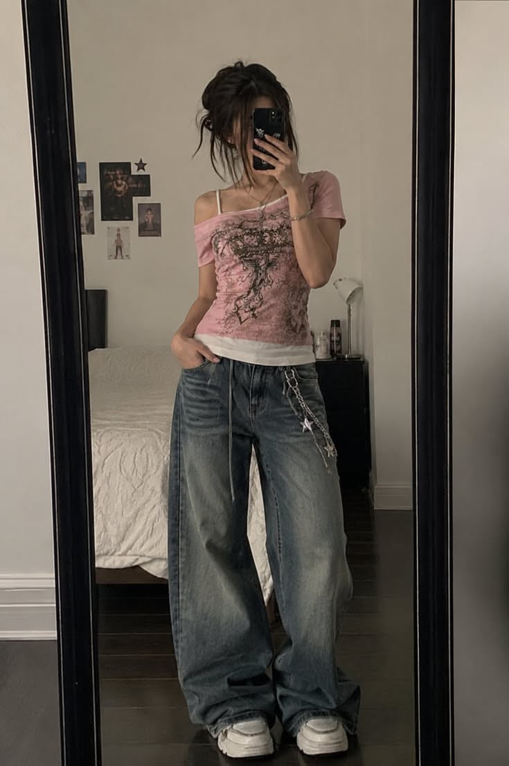 Person in baggy jeans and a pink top taking a mirror selfie indoors