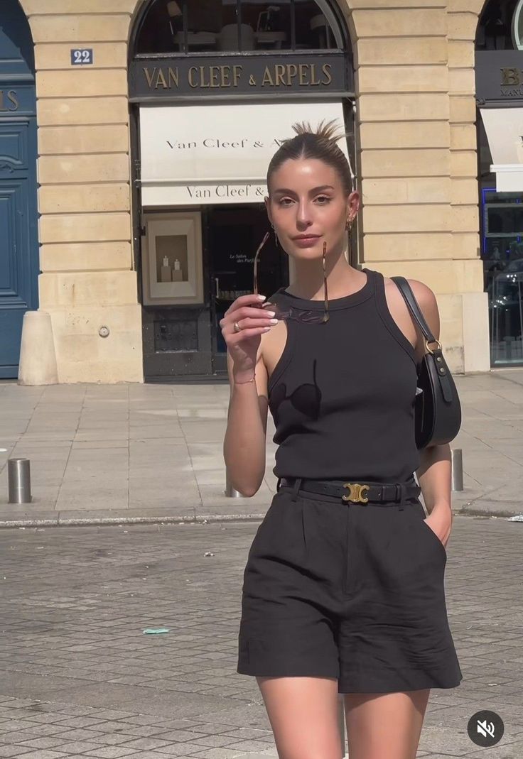 Woman in a black tank top and shorts holding sunglasses on a street