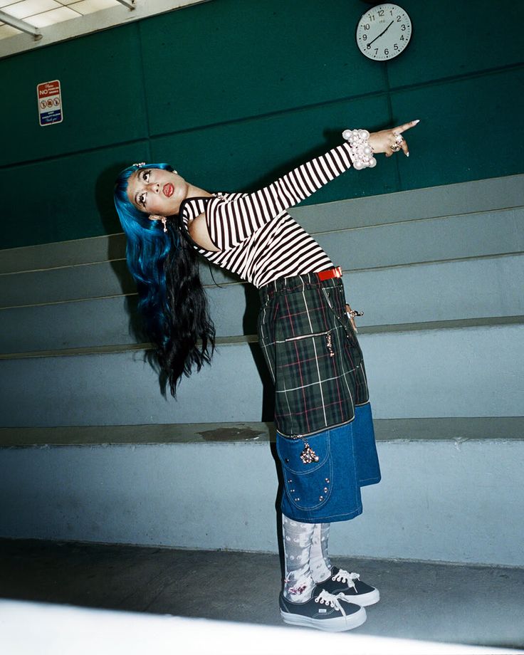 Woman with blue hair in a striped shirt and plaid skirt posing indoors