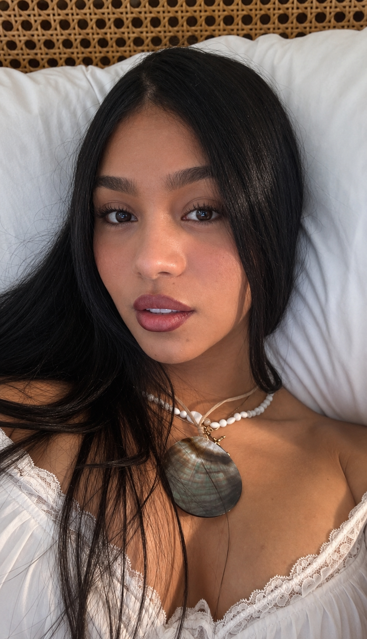 Woman with long black hair wearing a white top and shell necklace, lying on a pillow