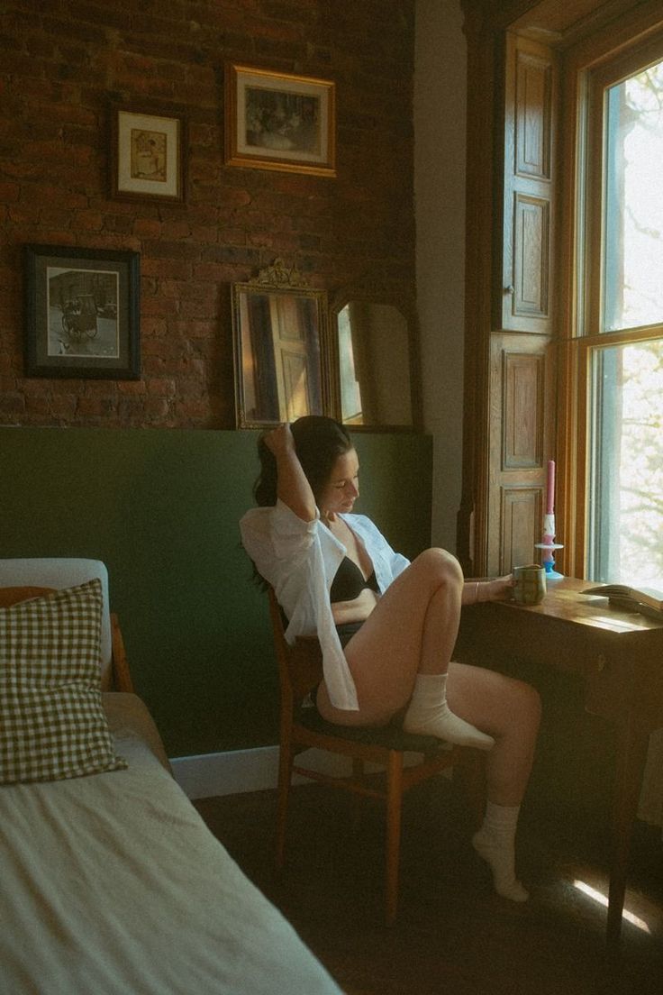 Woman in a bra and open white shirt sitting by a window in a warm-toned room