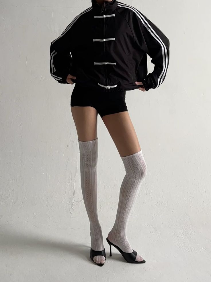 Person in a black jacket, short shorts, and thigh-high socks against a white wall