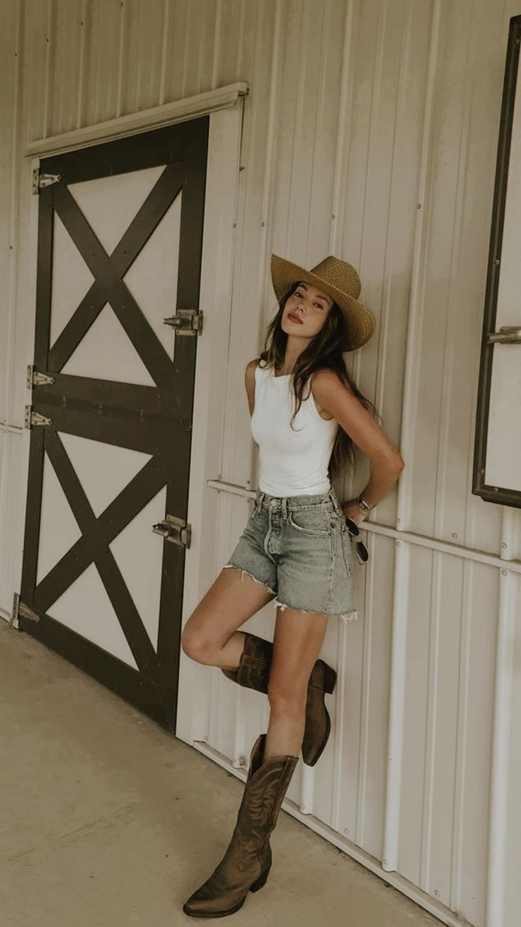 Woman in a cowboy hat, denim shorts, and cowboy boots leaning against a wall