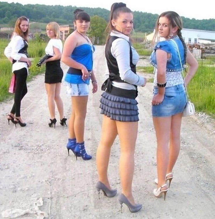 Five women in various outfits and high heels on a dirt road in the countryside
