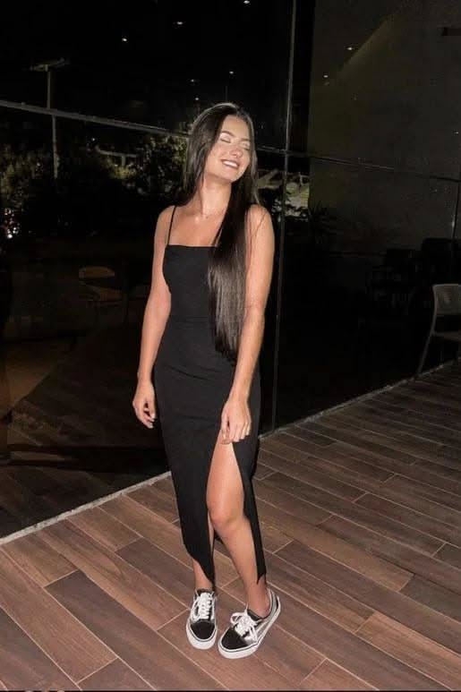 Woman in a black dress with a high slit and sneakers, smiling indoors at night