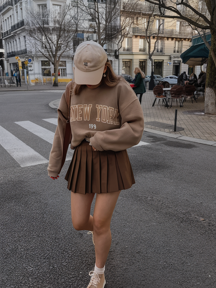 Person in a brown sweatshirt and pleated skirt walking on a street