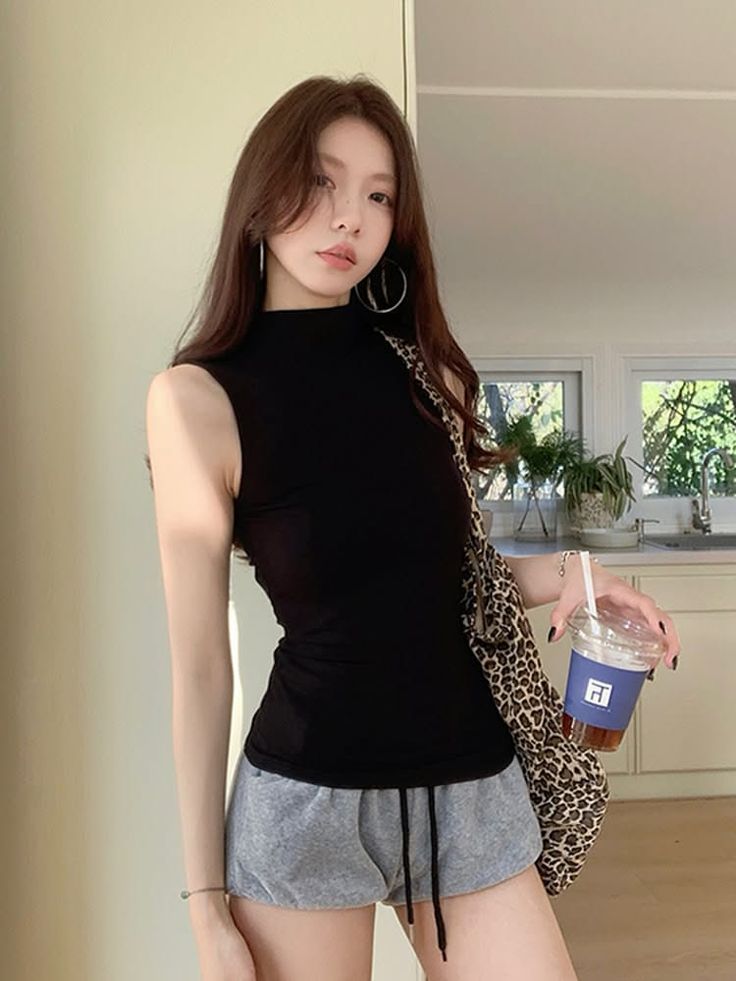 Woman with long hair in a black top and grey shorts holding a drink indoors