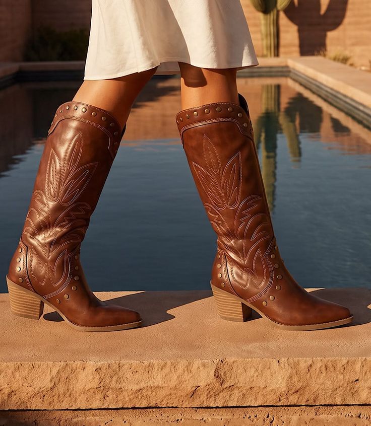Person wearing brown cowboy boots and a white dress by a pool in a desert setting