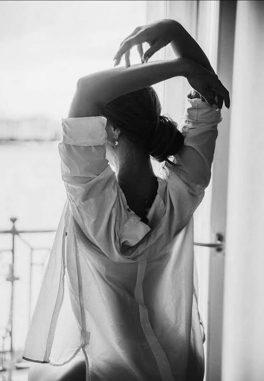 Woman in a loose shirt with arms raised, seen from behind in soft backlight